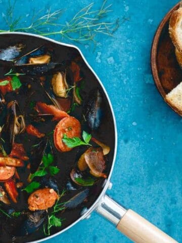 A pan of seafood stew with mussels, shrimp, and herbs sits on a blue surface beside a plate of sliced bread.