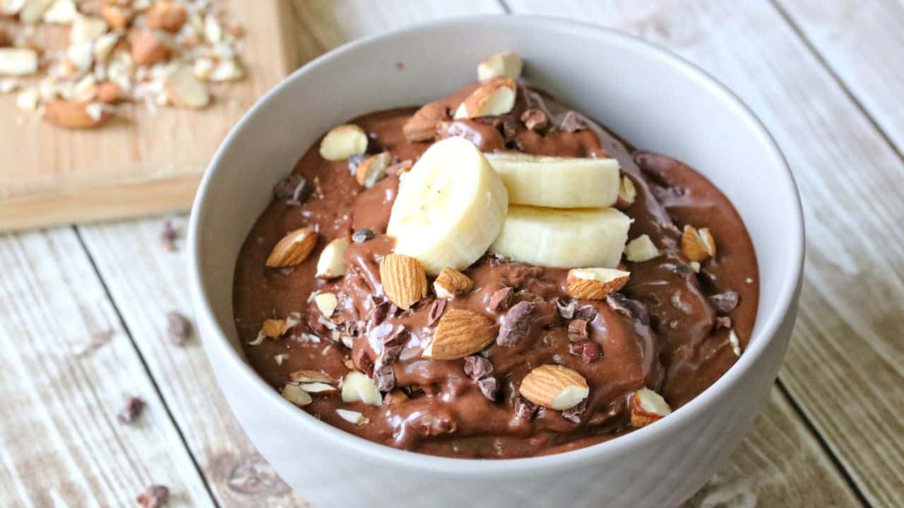 A bowl of chocolate smoothie topped with banana slices, chopped almonds, and cocoa nibs sits on a wooden table next to a cutting board with more nuts.