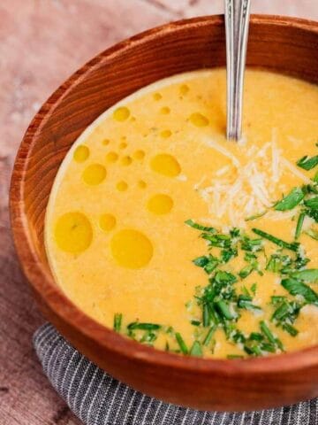 A bowl of creamy soup garnished with chopped herbs, grated cheese, and drizzled oil sits on a wooden surface next to a garlic bulb.