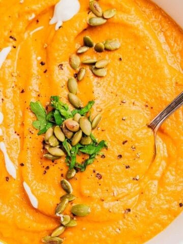 A bowl of orange-colored soup garnished with pumpkin seeds, chopped herbs, black pepper, and a drizzle of cream, with a spoon in the bowl.
