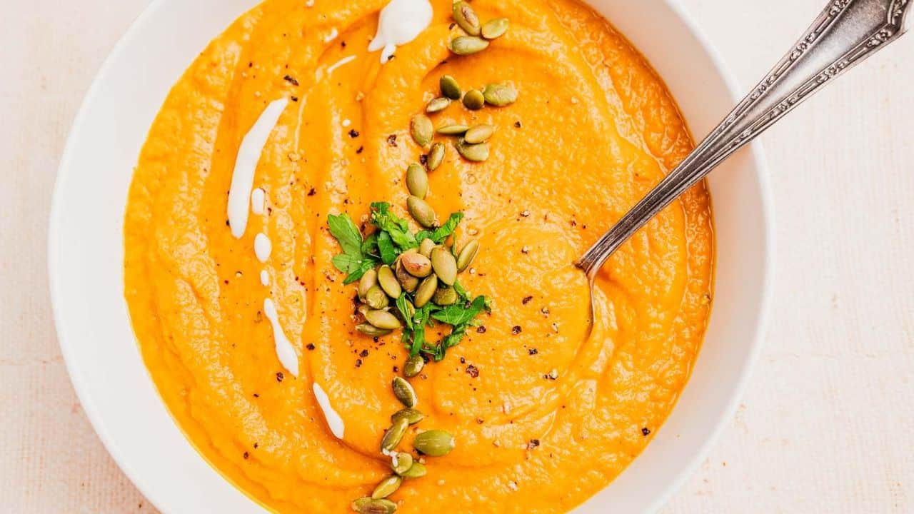 A bowl of orange-colored soup garnished with pumpkin seeds, chopped herbs, black pepper, and a drizzle of cream, with a spoon in the bowl.