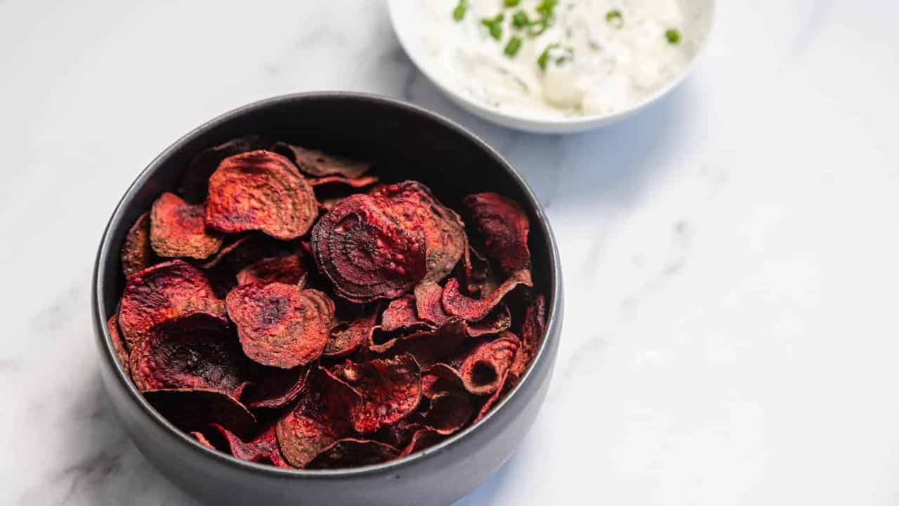 A black bowl filled with dark red beet chips sits on a white surface. In the background, there is a small white bowl containing a creamy dip with green herbs.