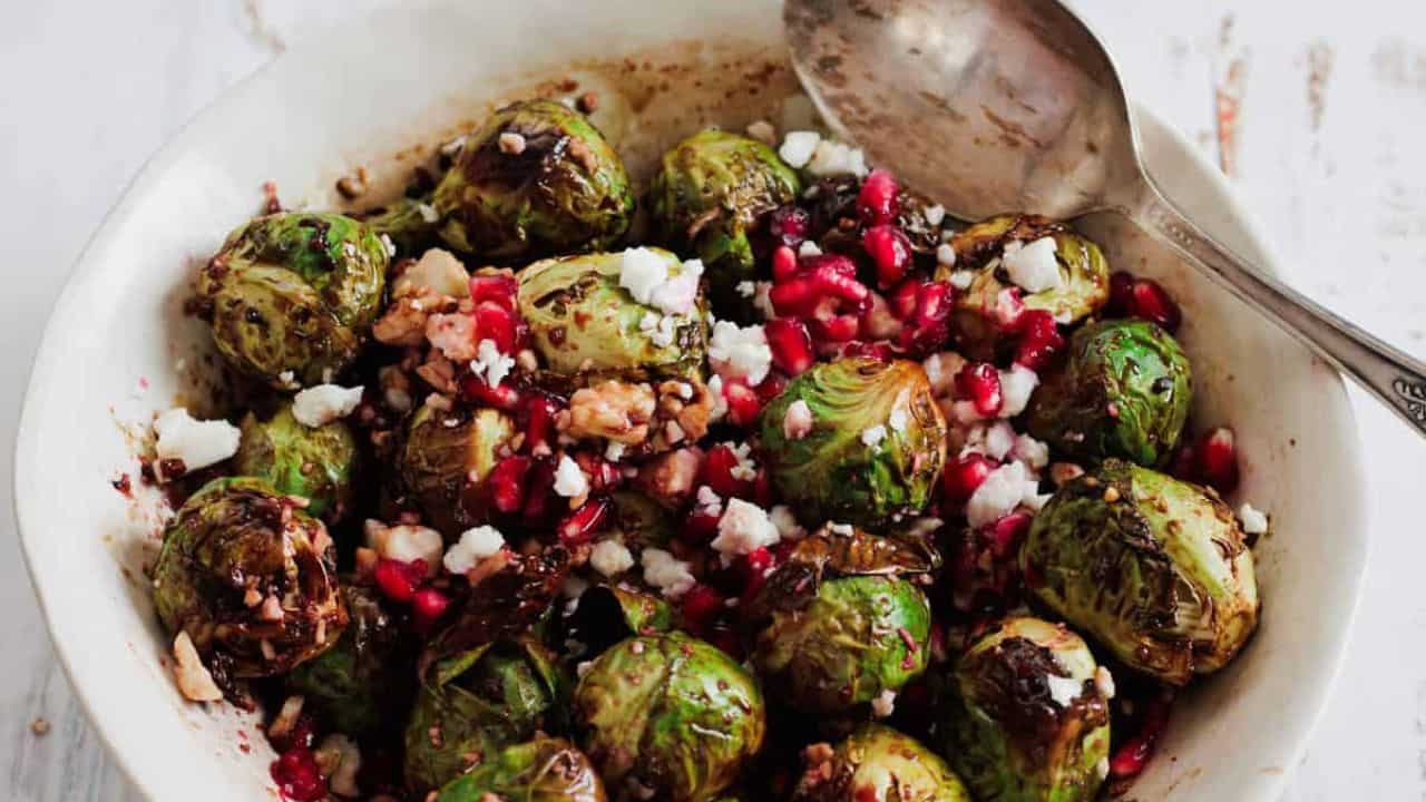 A wooden bowl filled with roasted Brussels sprouts. A jar of red sauce and a pair of tongs are in the background.