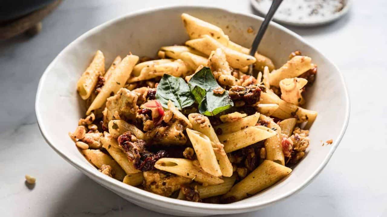 A white bowl filled with penne pasta mixed with chunks of chicken, sun-dried tomatoes, and garnished with fresh basil leaves. A fork is placed in the bowl, and a small dish is visible in the background.