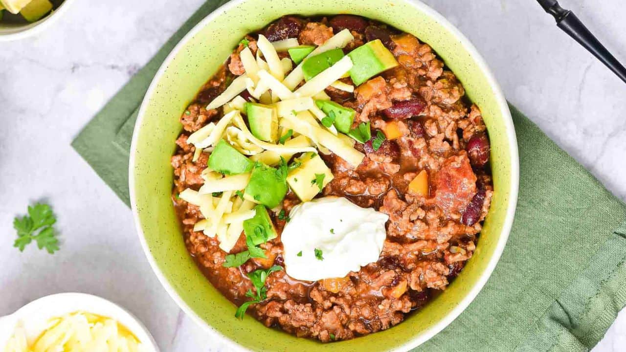 A bowl of chili topped with shredded cheese, diced avocado, and a dollop of sour cream, with a fork resting in the bowl. The food is served in a light green bowl on a green napkin.