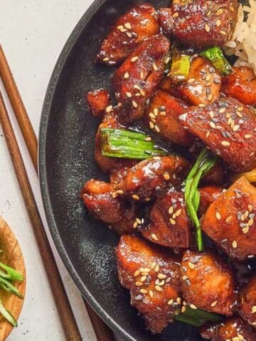 A plate of glazed chicken pieces topped with sesame seeds and green onions, served with a portion of white rice. Chopped green onions, sesame seeds, and chopsticks are nearby.