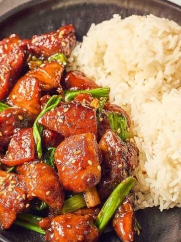 A black plate with two scoops of white rice and a serving of glazed chicken with green onions and sesame seeds, next to a glass of amber-colored beverage.