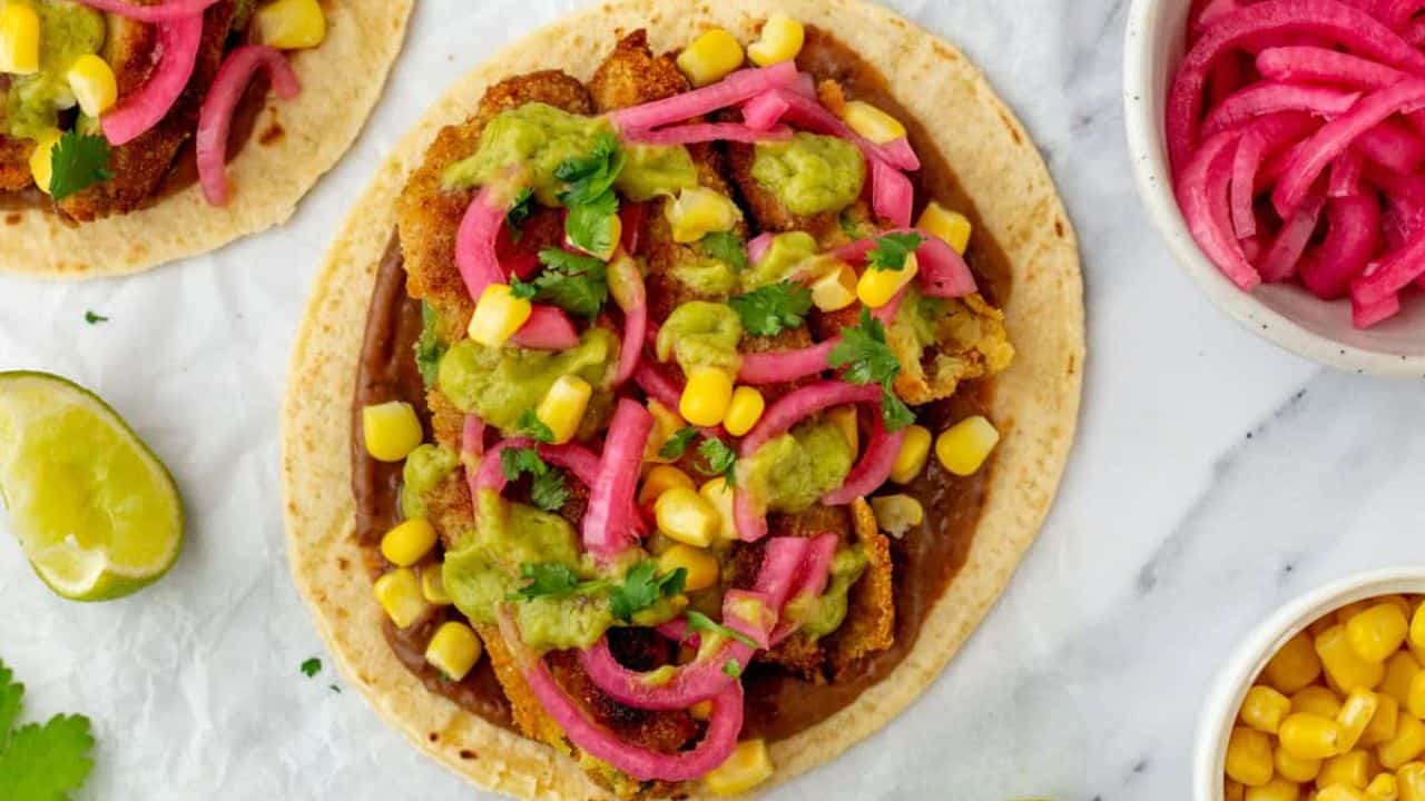 An overhead view of three fried avocado tacos on parchment paper surrounded by garnishes.