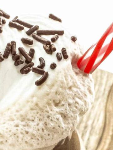 A milkshake topped with whipped cream, chocolate sprinkles, a maraschino cherry, and a red-striped straw on a wooden surface.