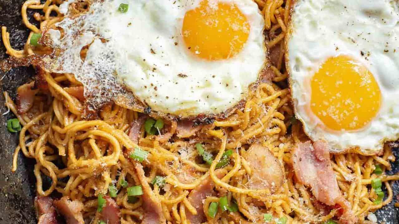 A person uses chopsticks to lift a portion of Gochujang noodles with crispy bacon and two sunny-side-up eggs from a skillet.