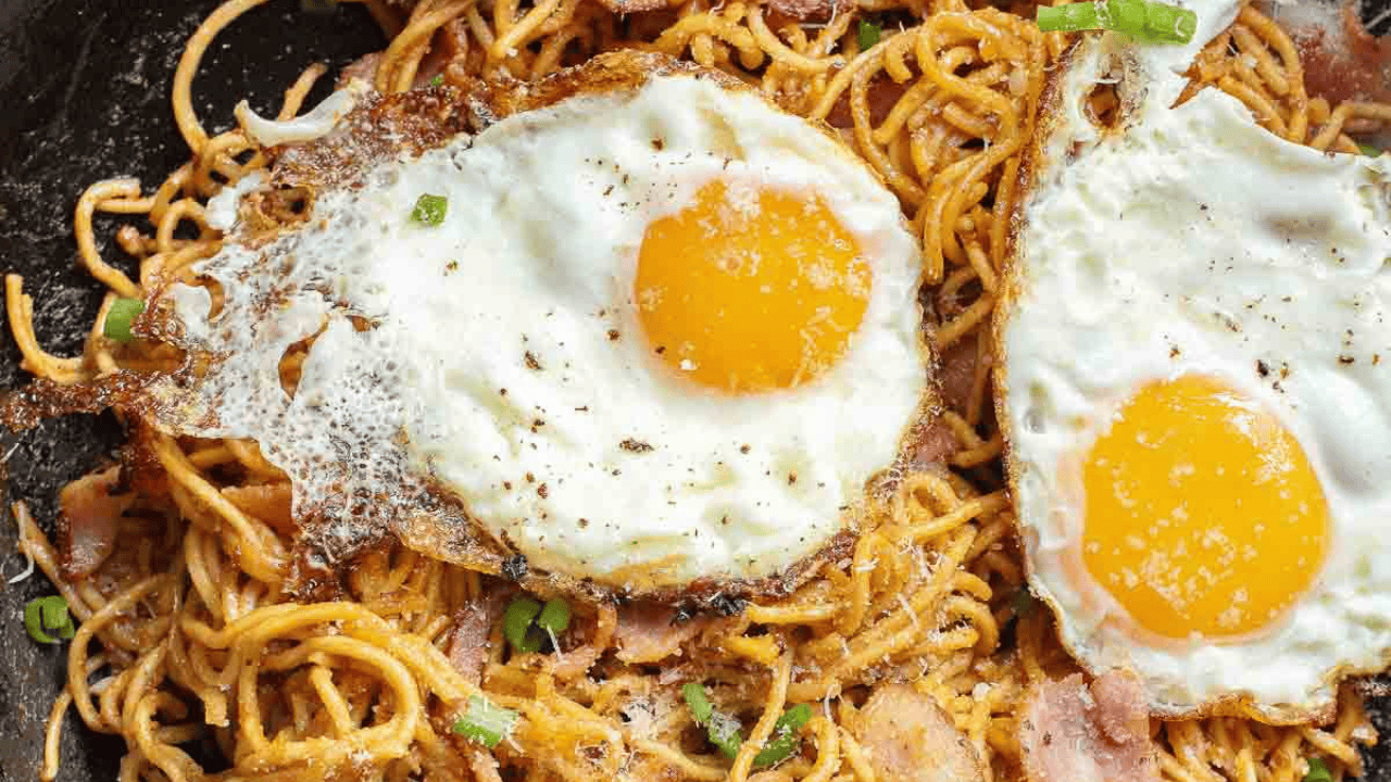 A person uses chopsticks to lift a portion of Gochujang noodles with crispy bacon and two sunny-side-up eggs from a skillet.