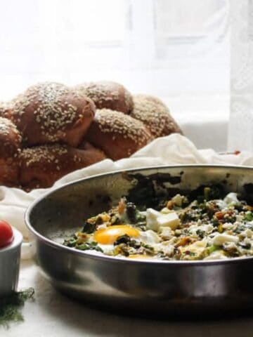 A table set with a skillet of baked eggs and greens, sesame bread, fresh vegetables, cubes of cheese, and a glass of tea in front of a window with sheer curtains.
