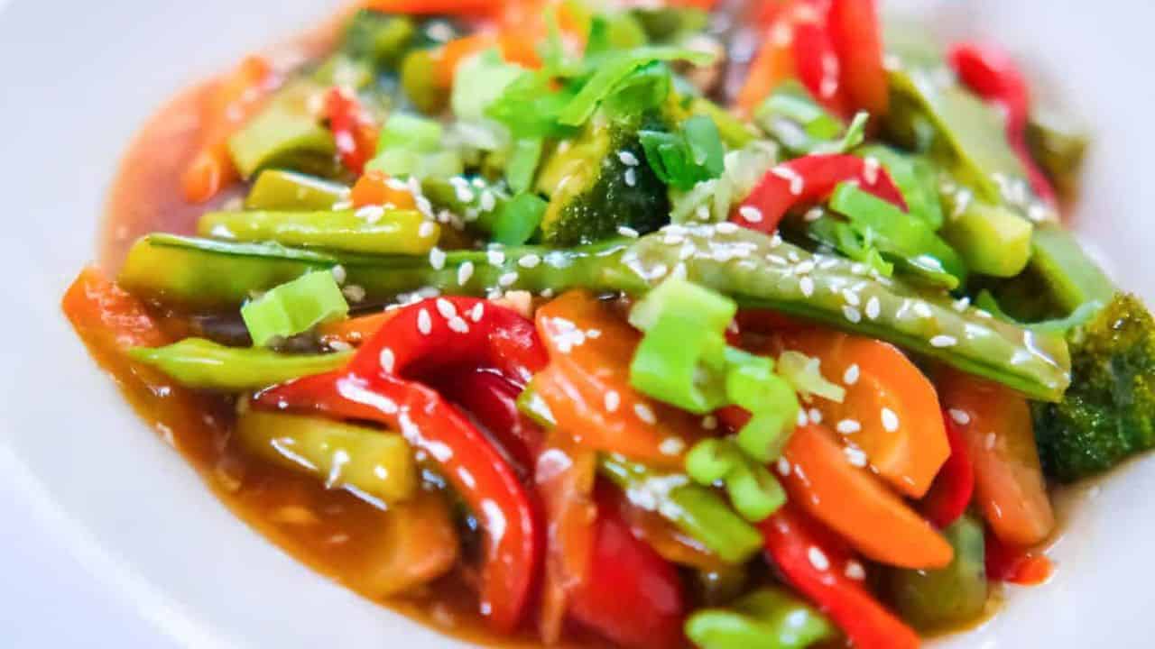 Veggie teriyaki stir fry with broccoli, carrots, red bell peppers, and snap peas in a savory sauce, topped with sesame seeds and green onions in a white bowl.