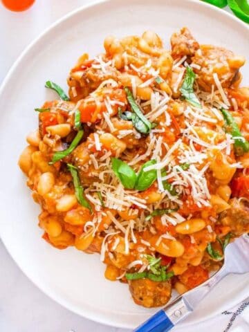 A plate of pasta with tomato sauce, white beans, grated cheese, and fresh basil, with a skillet of the same dish and fresh herbs nearby.