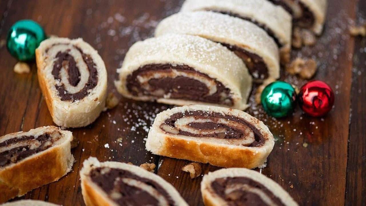 Sliced nut roll pastries with a chocolate swirl filling are arranged on a wooden surface, with powdered sugar sprinkled on top and two Christmas ornaments nearby.