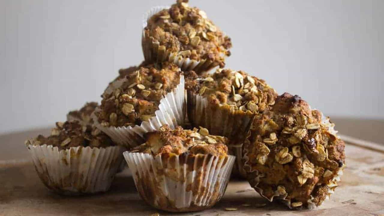 A pile of oat muffins in white paper liners is arranged on a wooden surface against a plain background.