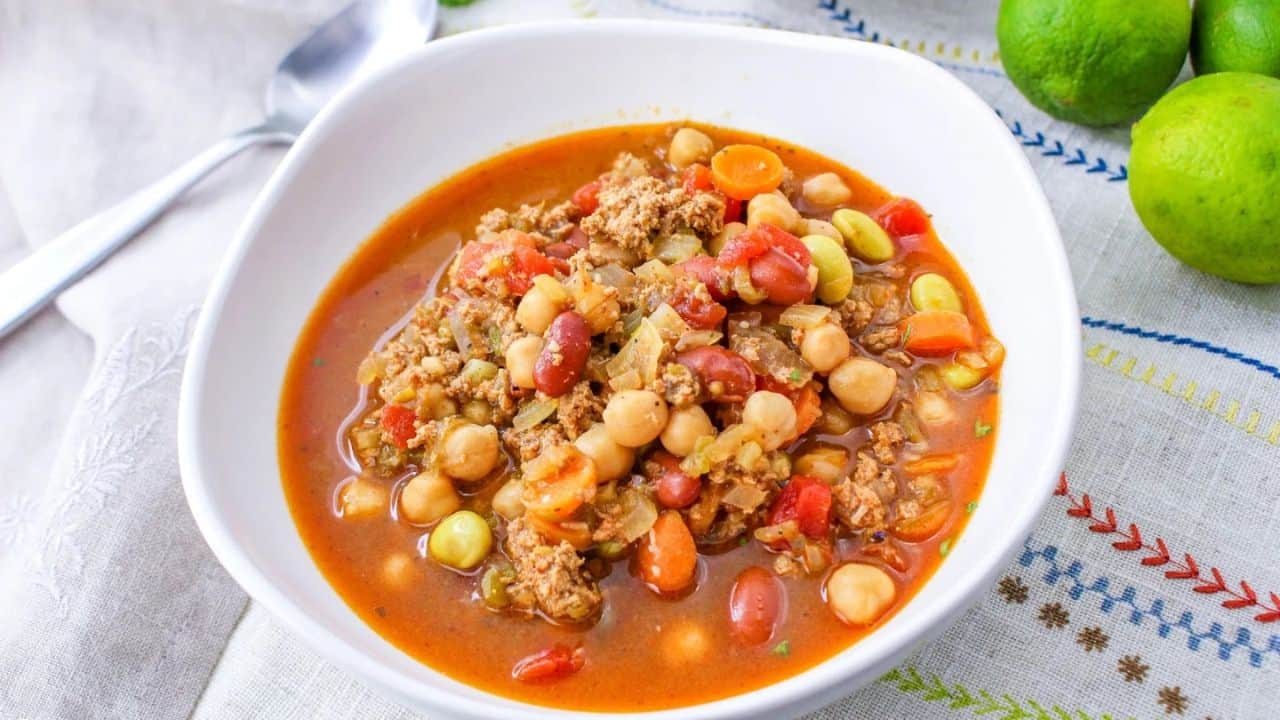 A bowl of colorful bean and vegetable chili, featuring chickpeas, red kidney beans, carrots, and ground meat, placed on a patterned cloth with limes and a spoon nearby.