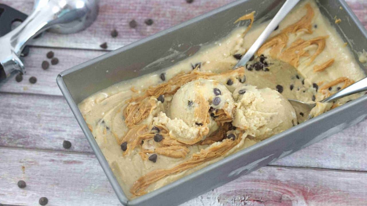 A metal loaf pan filled with homemade ice cream, swirled with peanut butter and chocolate chips, with two metal scoops inside.