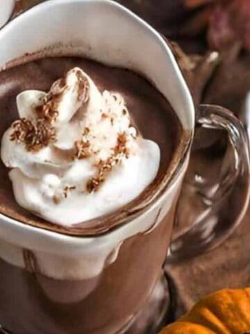 A clear mug of hot chocolate topped with whipped cream sits on a wooden surface, surrounded by a small pumpkin and autumn-themed decor.