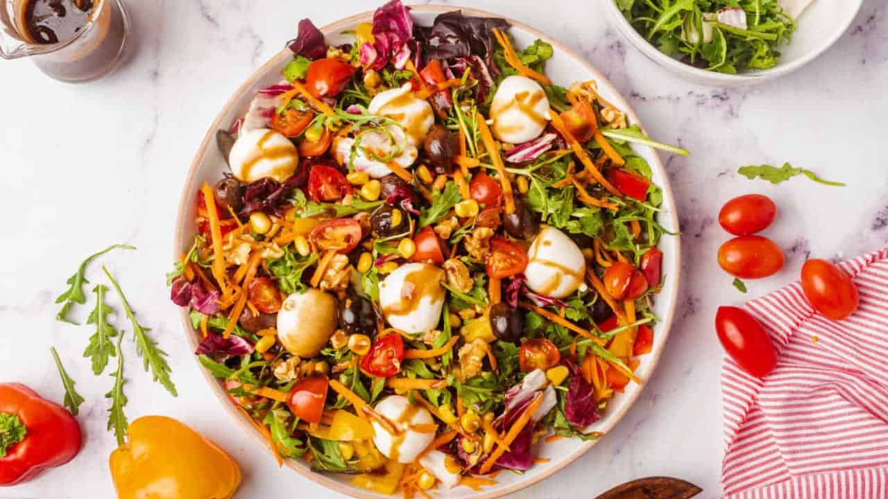 A rainbow salad with lettuce, tomatoes, carrots, olives, corn, and mozzarella balls is drizzled with dressing.