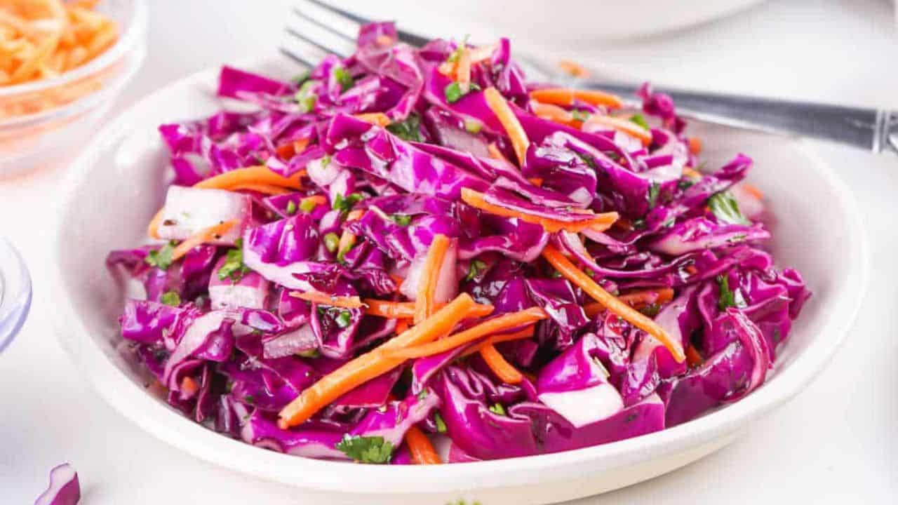 A bowl of red cabbage slaw with shredded carrots and chopped herbs, served with a fork on the side.