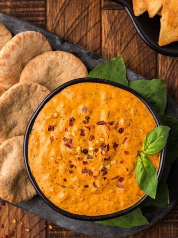 A black platter with round pita bread slices arranged around a bowl of orange dip, garnished with red pepper flakes and basil, sits on a wooden table next to a striped cloth.