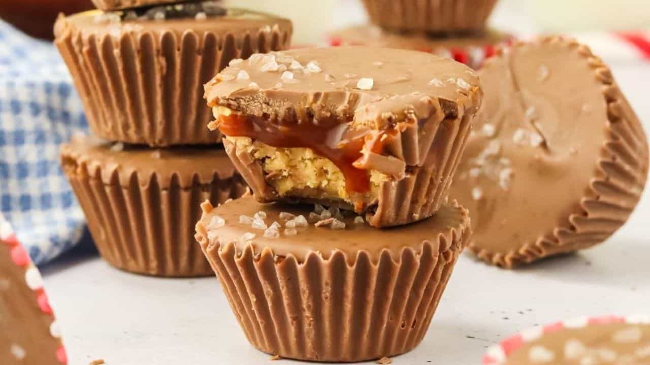 Chocolate cups stacked and arranged, with one cut open to show caramel and cookie layers inside, topped with coarse sea salt.