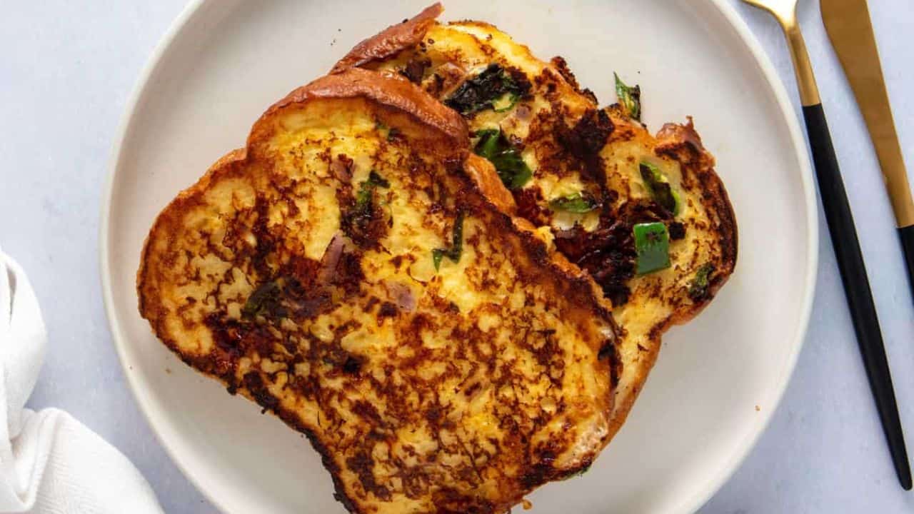 Close-up image of savory bombay toast on a white plate, with a fork and knife placed beside the plate.