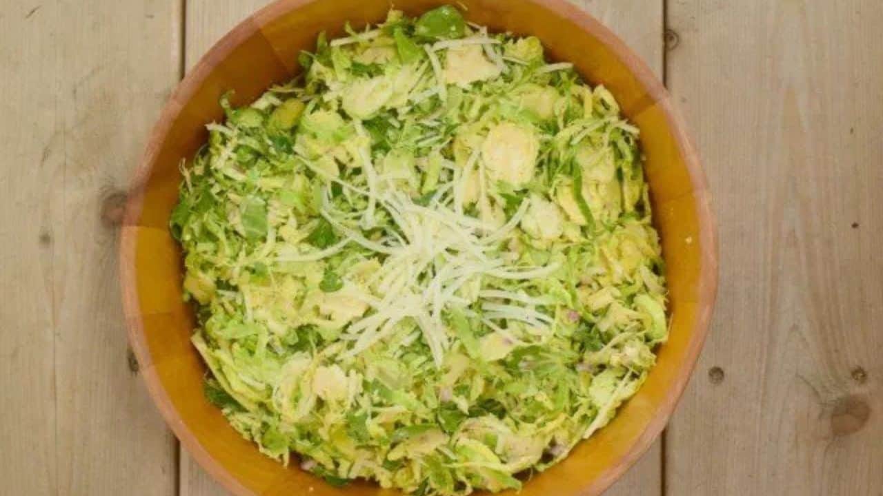 Image shows a fork holding a bite of shaved brussels sprouts salad with a clear bowl containing the rest of the serving behind it.