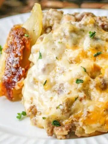 A serving of cheesy, baked casserole with ground meat and potatoes on a white plate, garnished with chopped herbs, next to a fork.