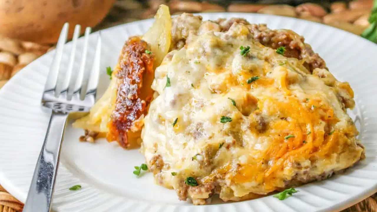 A plate of cheesy beef and potato casserole garnished with herbs, served with a fork. A potato, greenery, and a slow cooker are visible in the background.
