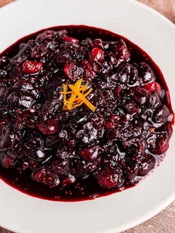 A white plate filled with cranberry sauce garnished with orange zest, placed on a wooden table with a spoon, cinnamon sticks, star anise, and a small bowl of cranberries nearby.