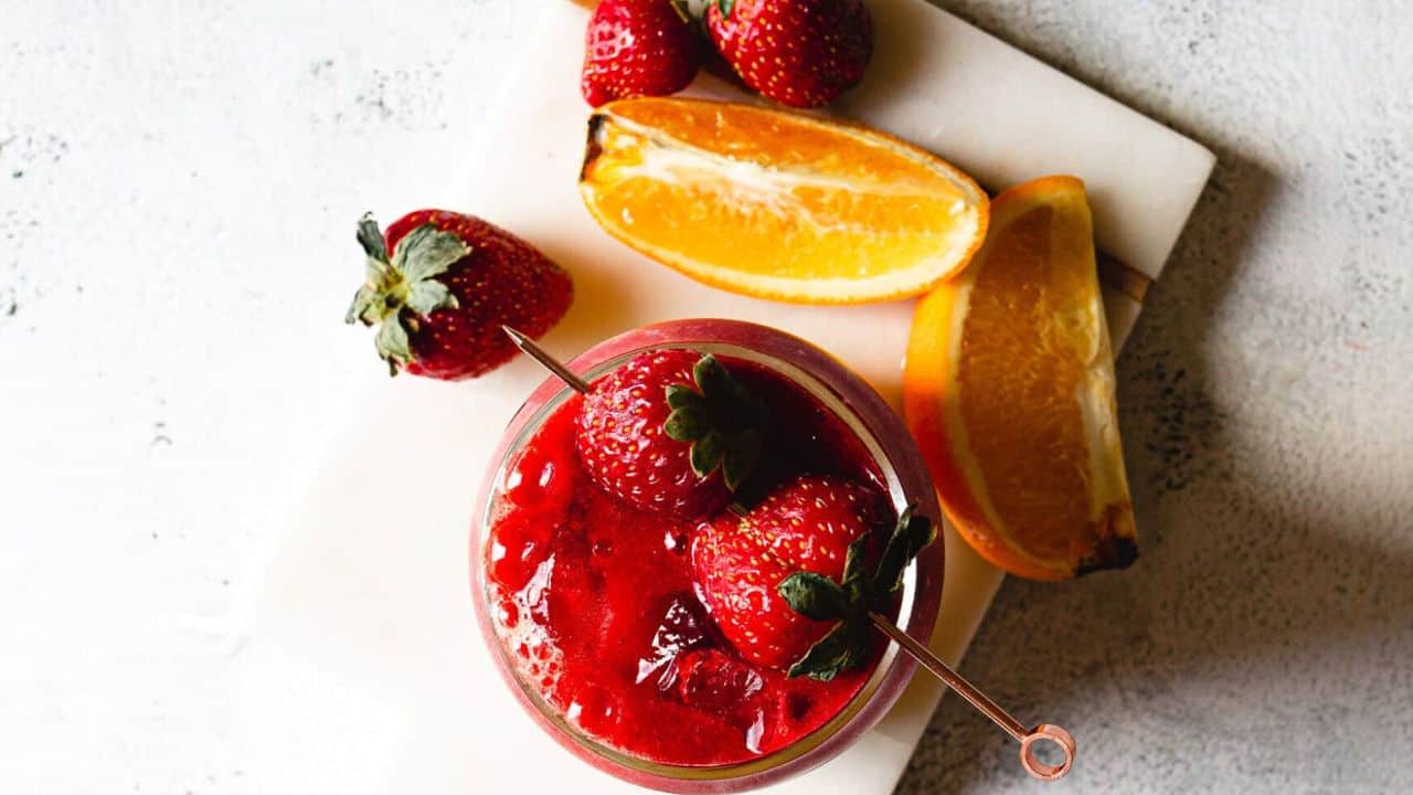 A glass filled with a red slushy drink, topped with whole strawberries on a skewer. The surface below is light and slightly textured, contrasting with the dark background.