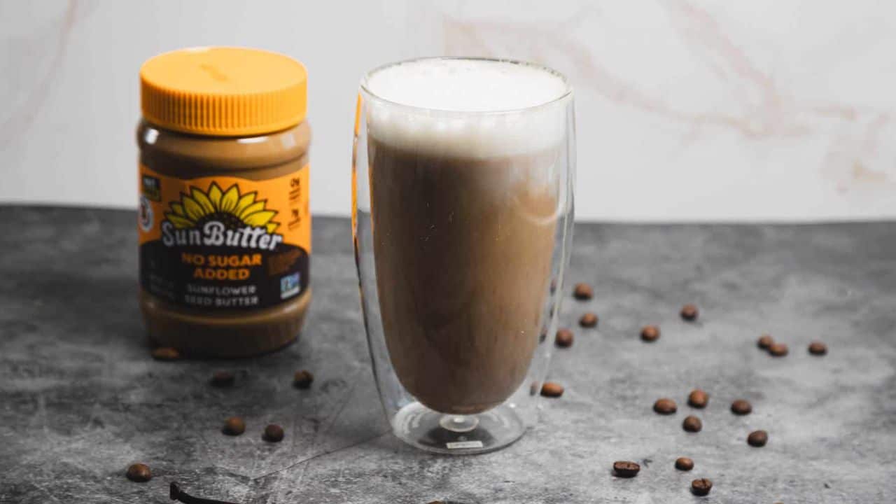 A glass of iced coffee with foam on top sits on a dark surface, surrounded by scattered coffee beans. A jar of SunButter 