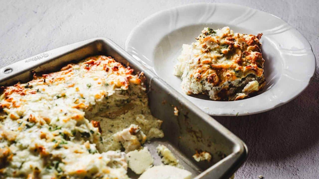 A person is using a spatula to serve a portion of baked mashed potato casserole from a metal baking dish. The casserole is topped with melted cheese and sprinkled with herbs.