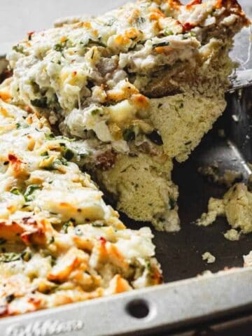 A metal baking dish with a cheesy casserole being sliced and served using a spatula.