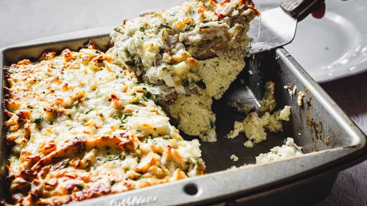 A person is using a spatula to serve a portion of baked mashed potato casserole from a metal baking dish. The casserole is topped with melted cheese and sprinkled with herbs.