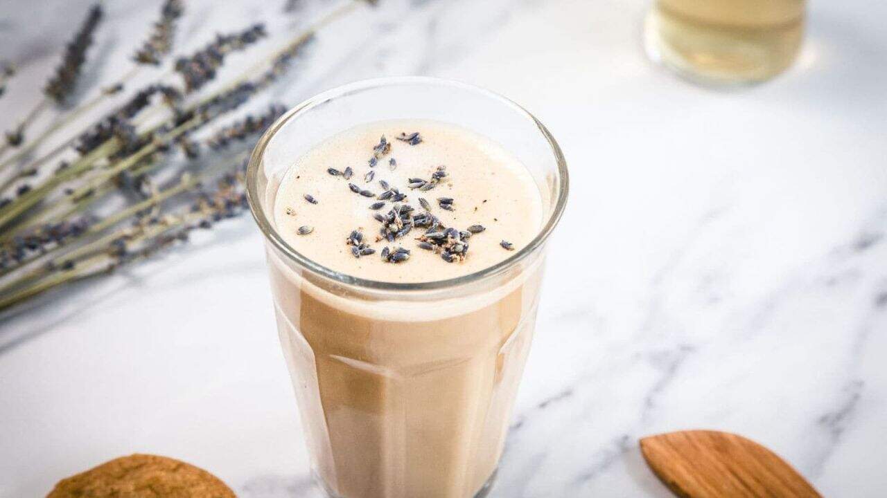 A glass of frothy iced coffee topped with dried lavender buds sits on a white marble surface. Lavender sprigs, a wooden spoon, and a cookie are nearby.