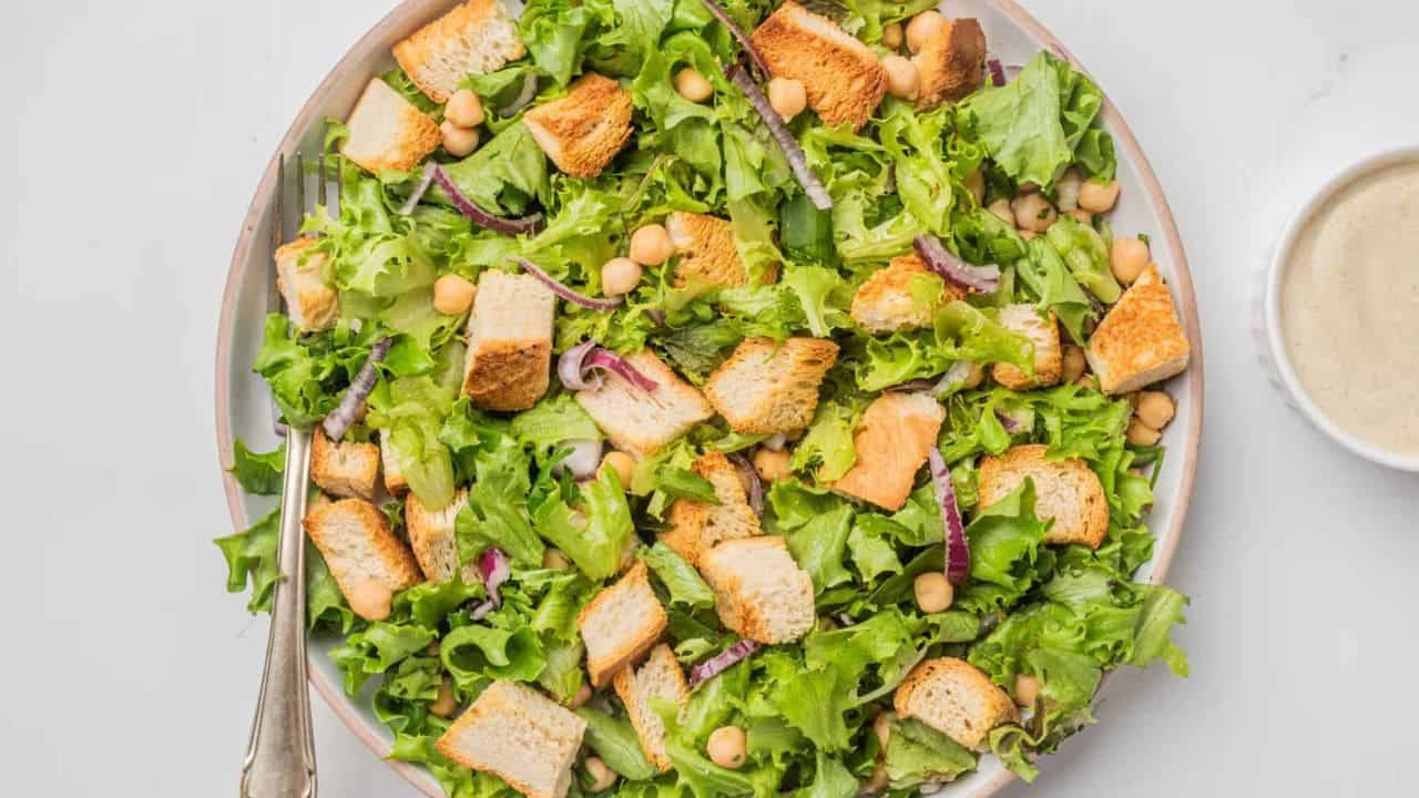 A plate of green salad with lettuce, croutons, chickpeas, and red onion slices, served with a fork and a small bowl of dressing on the side.