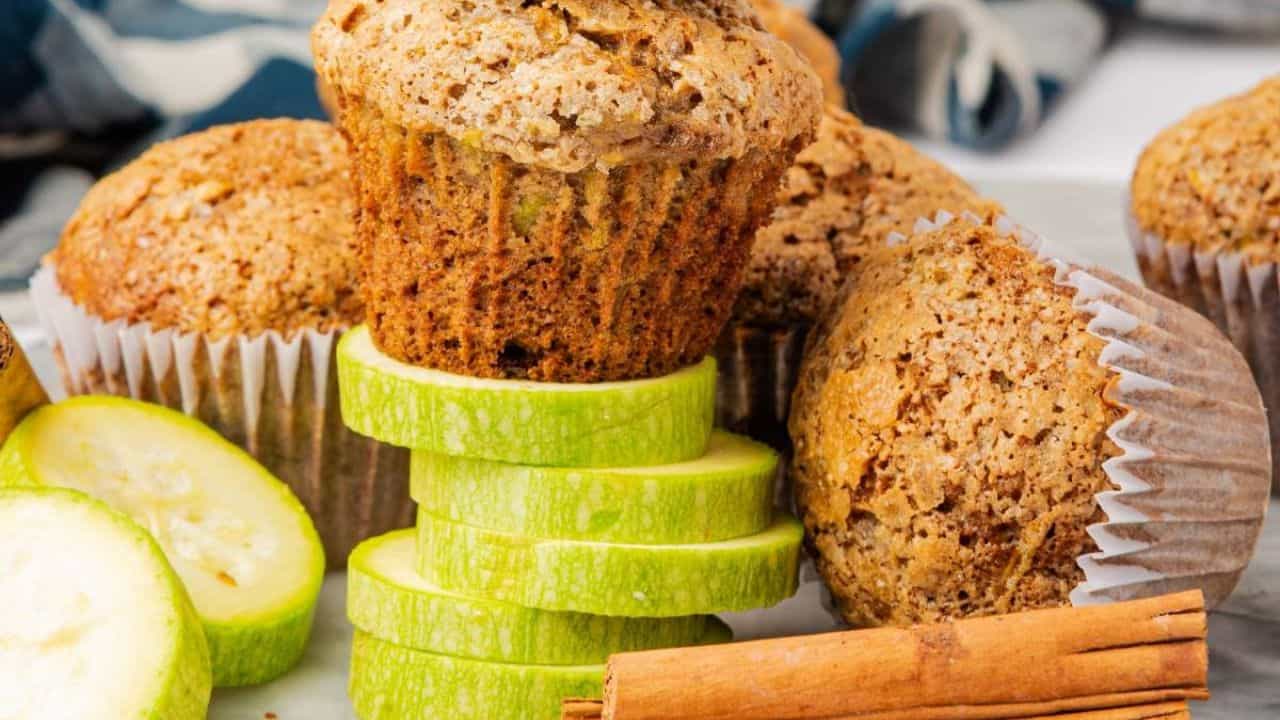 A stack of sliced zucchini sits in front of several zucchini muffins in paper liners, with a cinnamon stick placed nearby.