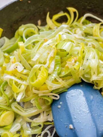 Sliced leeks being sautéed in a skillet with a blue spatula and pieces of garlic.