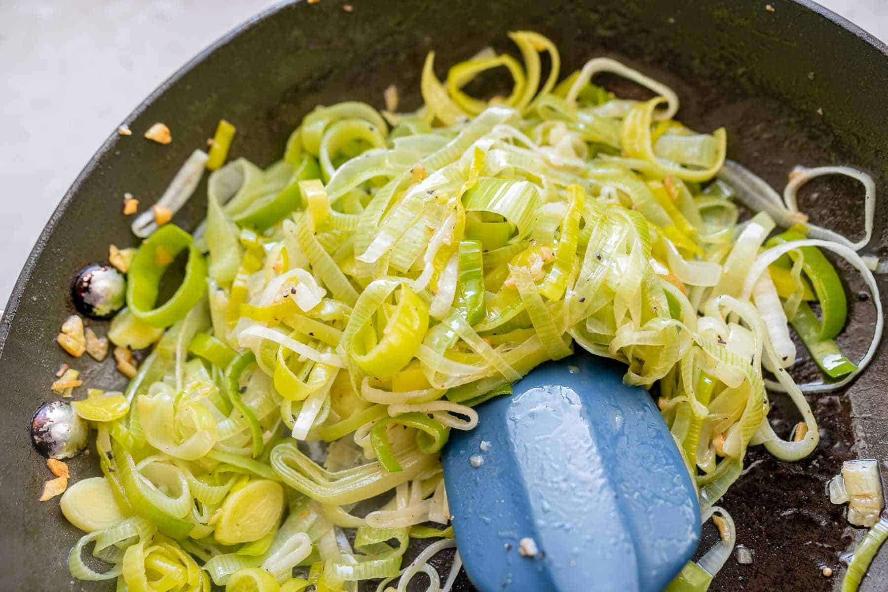 Sliced leeks being sautéed in a skillet with a blue spatula and pieces of garlic.