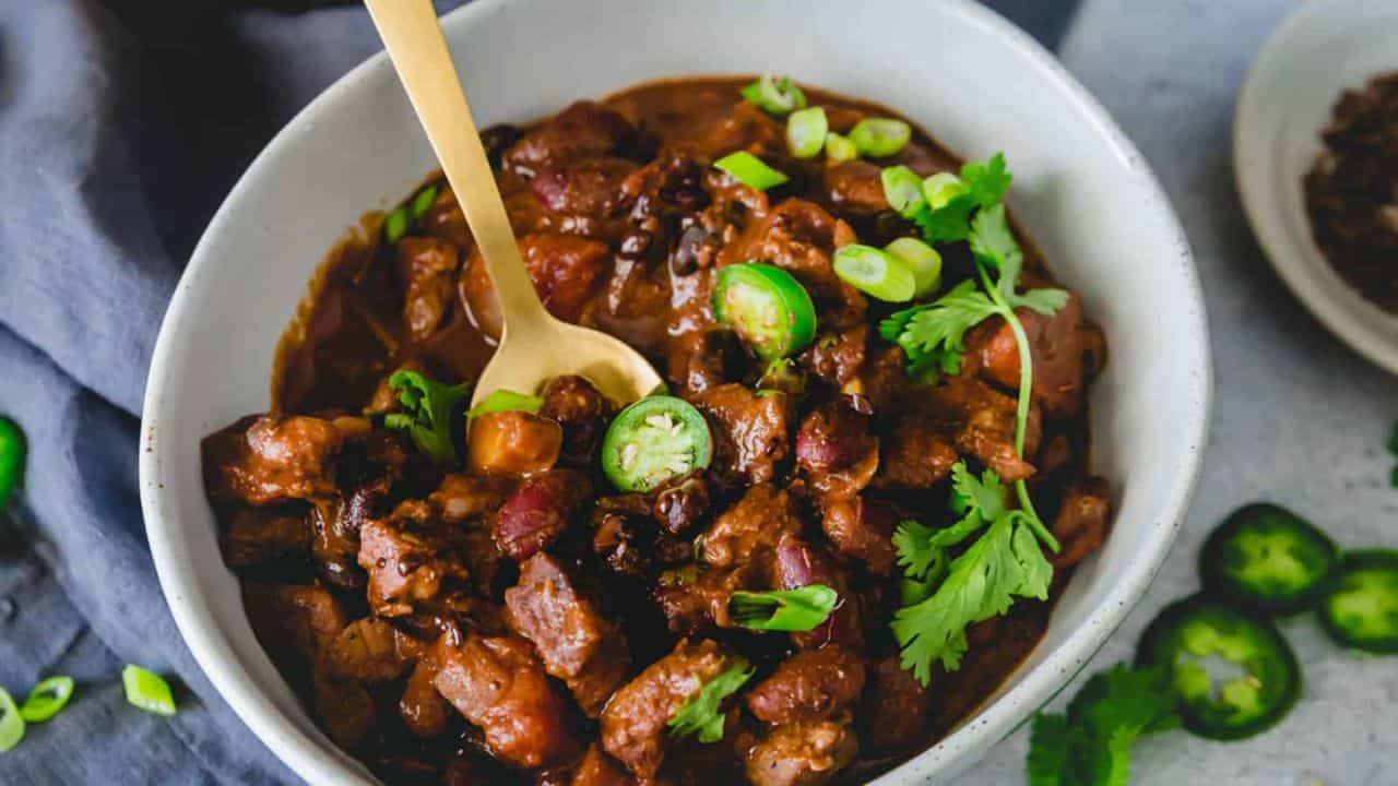 A bowl of lamb chili garnished with cilantro, jalapeños and green onions with a gold spoon.