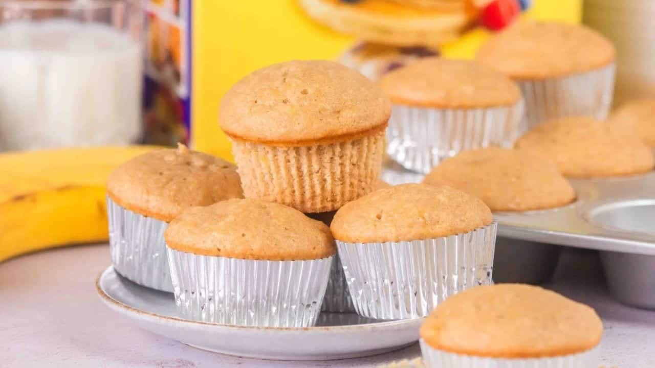 A plate of banana muffins in silver paper liners is displayed, with additional muffins in a muffin tin and a banana in the background.