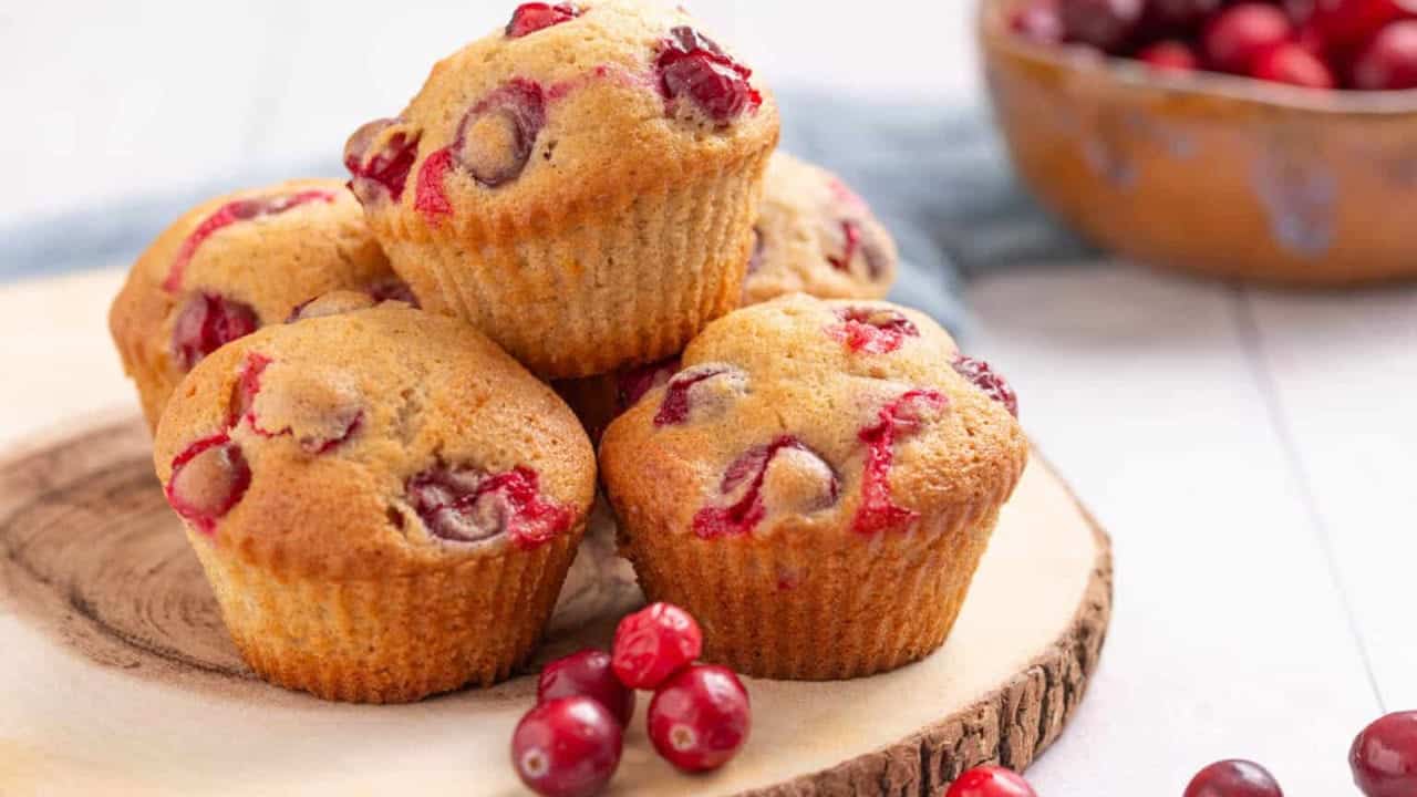 Four cranberry muffins are stacked on a wooden plate, with fresh cranberries placed around them. A bowl of cranberries is visible in the background.