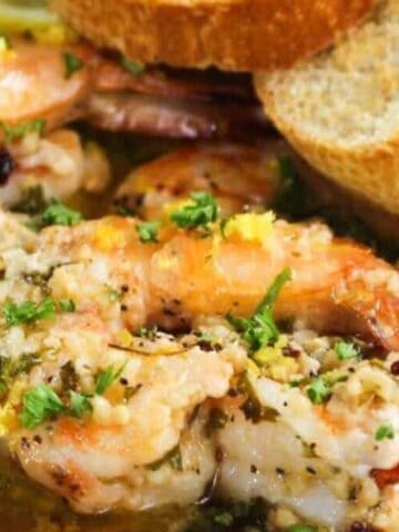 Two cooked shrimp garnished with herbs and garlic sit in a buttery sauce on a plate next to several slices of toasted bread.
