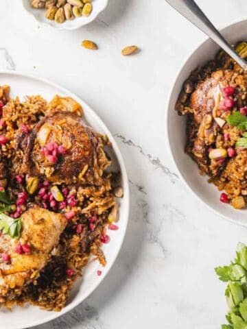 Two white bowls filled with spiced rice and chicken, garnished with pomegranate seeds, pistachios, and parsley, placed on a marble surface with fresh herbs nearby.
