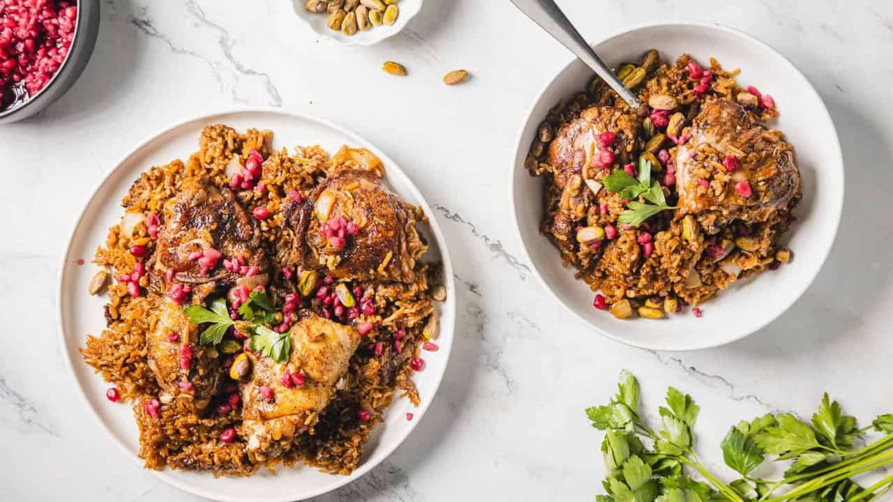 A close-up of a dish featuring seasoned rice topped with cooked chicken pieces. The meal is garnished with fresh parsley, pomegranate seeds, and pistachios, served on a white plate.