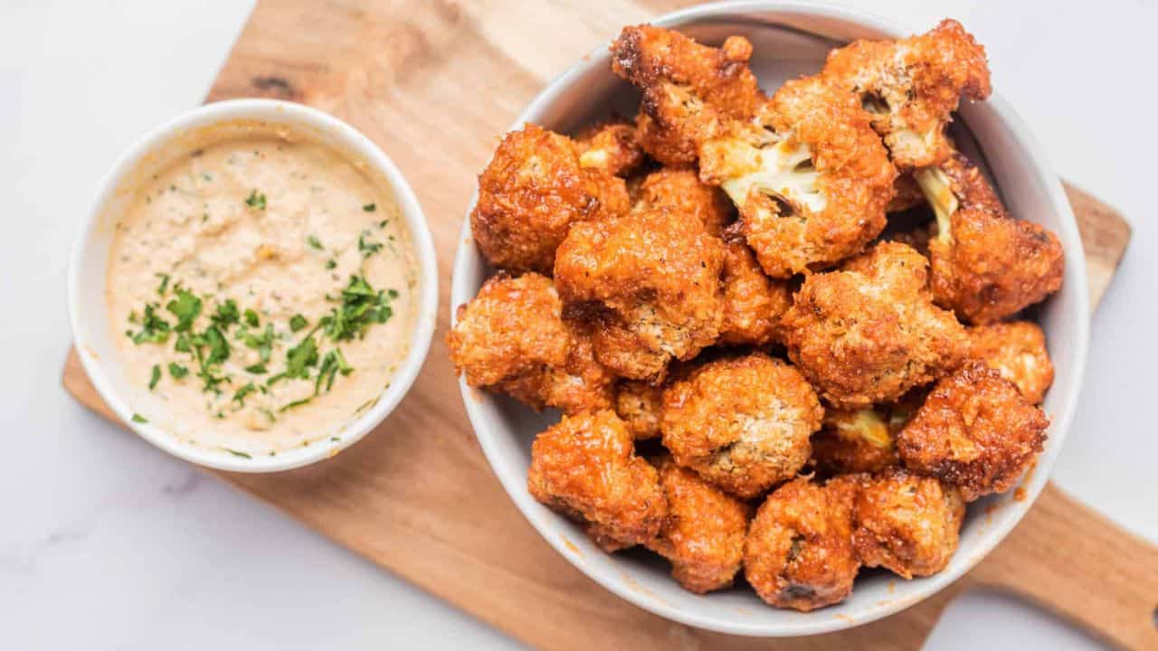 Air fryer vegan buffalo cauliflower bites in a bowl with dip nearby, placed under a wooden board.
