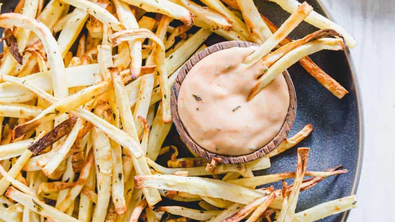 Parsnip fries on a plate with dipping sauce on a blue kitchen towel.